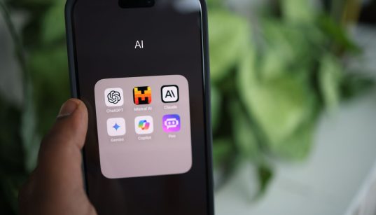 Mão segurando um smartphone com aplicativos de inteligência artificial exibidos na tela.