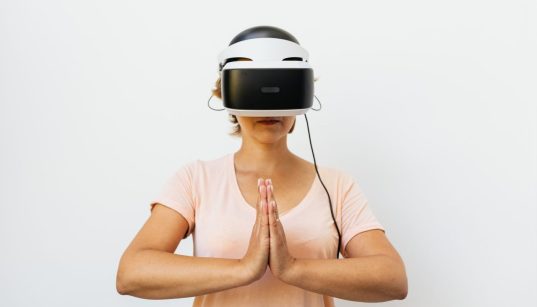 Mulher adulta medita usando um headset de realidade virtual diante de fundo branco.
