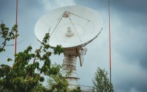 Grande antena parabólica de comunicação via satélite diante de um céu nublado, cercada por vegetação.