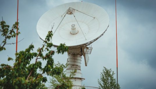 Grande antena parabólica de comunicação via satélite diante de um céu nublado, cercada por vegetação.