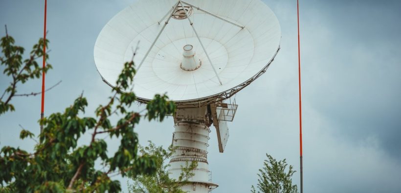 Grande antena parabólica de comunicação via satélite diante de um céu nublado, cercada por vegetação.