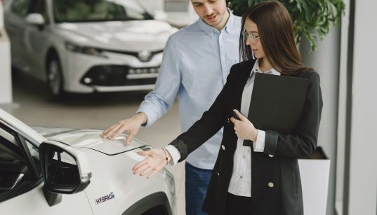 Homem conversa com uma vendedora sobre as características de um veículo híbrido em um showroom de automóveis.